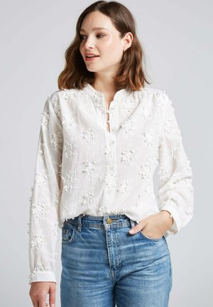 Blouse blanche à manches longues avec broderie florale texturée, patte de boutonnage et coupe décontractée, associée à un jean bleu.