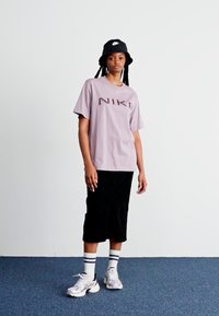 Camiseta Nike de color púrpura claro con logo marrón, falda midi negra, calcetines blancos con rayas y zapatillas grises. Diseño sencillo y corte relajado.