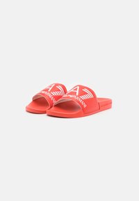 Sandálias de slide vermelhas feitas de borracha, com uma faixa larga com logotipo branco e design de asas, sola plana e textura suave.