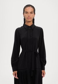 Melna kleita ar apkakles kaklu, garām piedurknēm un pogu aizdari. Iezīmē plānu svārku, teksturētas detaļas un detalizētu jostas saitīti.