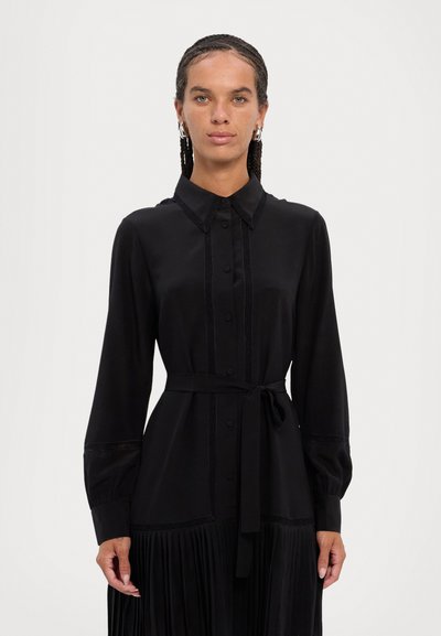 Vestido negro con cuello tipo camisa, mangas largas y cierre de botones. Presenta una falda plisada, detalles texturizados y un lazo en la cintura.