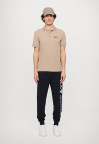 Beige polo skjorte med sorte detaljer og logo, kombinert med marineblå joggebukser med hvit tekst. Modellen har på seg en matchende beige caps og hvite sneakers.