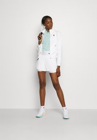 Hvid atletisk jakke med lynlås, lyseblå polo og hvide shorts. Båret med hvide og teal sneakers. Enkel design med sporty detaljer.