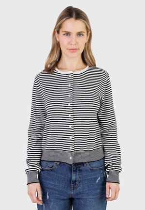 Junge Frau mit blondem Haar, die eine schwarz-weiß gestreifte, geknöpfte Strickjacke und blaue Jeans trägt, vor einem schlichten hellen Hintergrund stehend.