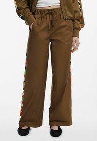 Hnedé široké nohavice s pásom na šnúrku, so farebnými vzorovanými bočnými panelmi. Vyrobené z hladkej látky, vhodné na každodenné nosenie.