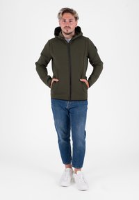 Giacca con cappuccio in tessuto verde oliva con zip frontale e tasche laterali, abbinata a jeans azzurri e sneakers bianche.