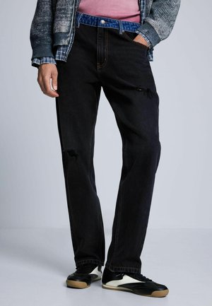 Persona che indossa jeans neri consumati, sneakers nere e bianche, una camicia rosa e maglioni blu e grigi a strati.