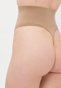 Primo piano di una persona che indossa un perizoma modellante beige a vita alta su uno sfondo bianco uniforme.