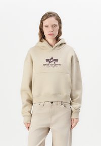 Persona che indossa una felpa beige di Alpha Industries e pantaloni beige chiaro abbinati, in piedi davanti a uno sfondo bianco.