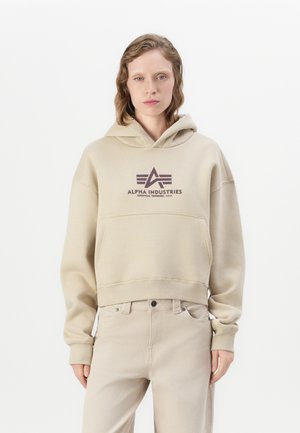Asmuo, vilkintis smėlio spalvos Alpha Industries gobtuvą ir derančias šviesiai smėlio spalvos kelnes, stovi prieš baltą foną.