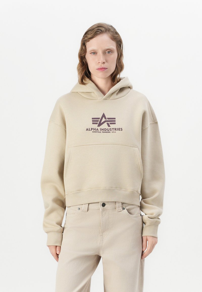 Persona vistiendo una sudadera beige de Alpha Industries y pantalones beige claros a juego, de pie contra un fondo blanco.