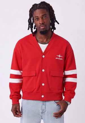 Homme avec dreadlocks porte un cardigan rouge boutonné avec des rayures blanches sur les manches, logo PXP sur la poitrine, jean bleu clair, et collier en chaîne argentée.