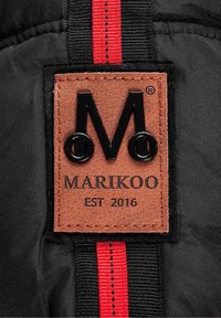 Écusson en cuir marron avec logo "M" noir, texte "MARIKOO" et "EST 2016" sur tissu noir avec rayure verticale rouge et noire.