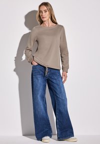 Felpa beige con scollo rotondo e orlo con cordino, abbinata a jeans in denim blu a gamba larga. Il modello si trova in piedi con le mani in tasca.