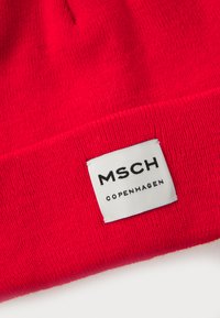 Red knitted beanie made of soft fabric, featuring a white label with "MSCH COPENHAGEN" printed in black. Close-up view shows texture.
