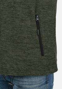 Suéter de forro polar verde con tejido texturizado, que presenta un bolsillo lateral con cremallera negra y un detalle de cordón en el dobladillo. Jeans de mezclilla azul visibles por debajo.