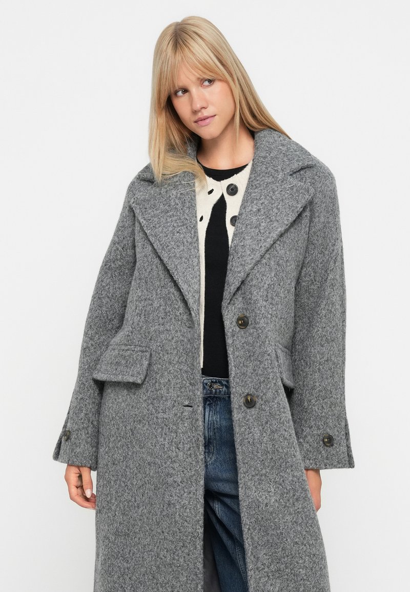 Manteau en laine gris avec un col à revers, de grands boutons, des poches latérales et une finition texturée. Coupe ample, design mi-long.