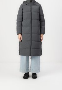 Lang grå pufferjakke med hætte, med quiltede sektioner, lynlås foran og sidelommer. Båret over lys blå brede jeans.