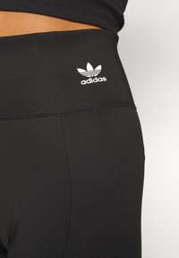 Melnas sporta legingi ar gludu tekstūru, aprīkoti ar plašu jostas daļu un baltu Adidas logo priekšā.