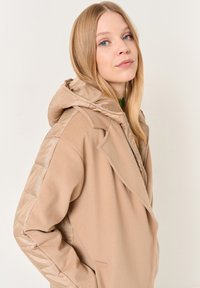 Giacca beige con un corpo in tessuto morbido e maniche lucide e imbottite. Caratterizzata da un cappuccio, chiusura con zip e tasche laterali.