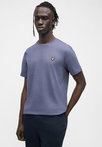 Hombre de piel oscura y cabello trenzado, vistiendo una camiseta azul de cuello redondo con un pequeño logo de un pájaro amarillo, de pie con una mano en el bolsillo.