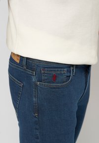 Modre denim hlače z ožjim krojem, ki imajo rdeč vezen logotip na zadnjem žepu in svetlo rjavo usnjeno etiketo na pasu.