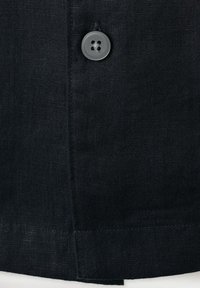 Detail černé látky s jediným černým čtyřděrovým knoflíkem přišitým poblíž svislého švu a lemu.