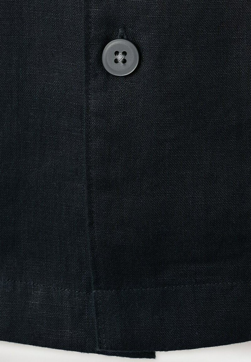 Gros plan sur un tissu noir avec un seul bouton noir à quatre trous cousu près d'une couture verticale et d'un ourlet.