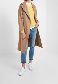 Cappotto di lana color cammello con design doppiopetto, abbinato a un maglione giallo, jeans azzurri chiari e sneaker bianche.