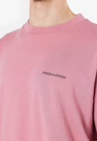 Tricou roz deschis din bumbac moale, cu guler rotund și un mic logo gri "PEGADOR" pe piept.
