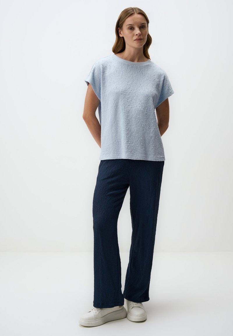 Lichtblauw textuur short-sleeve top gecombineerd met donkerblauw textuur wijde broek. Model draagt witte platform sneakers. Minimalistische achtergrond.