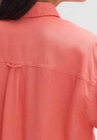 Korallenrotes Hemd mit weicher Textur, das einen Kragen und eine Kellerfalte auf der Rückseite aufweist. Feine Nähte verleihen dem Design zusätzlichen Akzent.