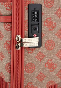 Valigia rigida con motivo rosso e zip dorate marchiate "GUESS?" e una serratura TSA nera su uno sfondo texture beige.