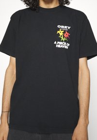Černé bavlněné tričko s krátkými rukávy. Na přední straně se nachází vícerozměrný grafický text "OBEY worldwide" a "A PIECE OF HEAVEN" s květinami.