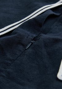 Gros plan d’un vêtement en tissu bleu marine foncé avec une fermeture éclair noire et deux bandes blanches parallèles le long de celle-ci.