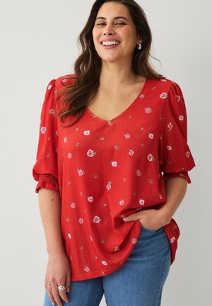 Smilende kvinde med langt brunt hår iført en rød blomstret bluse med pufærmer og blå jeans, med hånden i lommen, foran en lys baggrund.