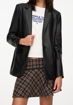 Femme portant un blazer en cuir synthétique noir, un t-shirt blanc avec un motif graphique et une jupe à carreaux marron avec un ourlet incliné sur fond uni.