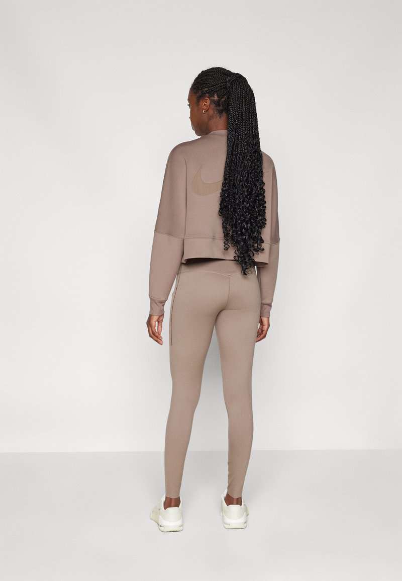 Bruine cropped sweatshirt en bijpassende leggings van een gladde stof. Bevat een tonale Nike-logo op de achterkant en witte sportschoenen.