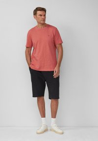 Rotes kurzärmeliges T-Shirt mit weicher Textur, kombiniert mit schwarzen knielangen Shorts und weißen Sneakern, steht vor einem grauen Hintergrund.