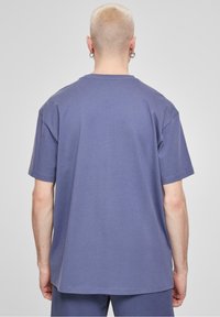 Persona con capelli corti decolorati che indossa una semplice maglietta viola oversize e pantaloni abbinati, in piedi con la schiena rivolta verso di noi su uno sfondo grigio.