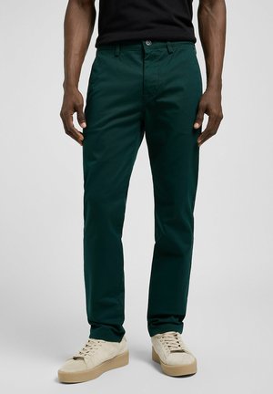 Dunkelgrüne Chinos aus glattem Stoff mit Knopfverschluss, Seitentaschen und gekrempelten Säumen. Kombiniert mit cremefarbenen Sneakers.