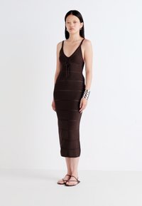 Modelis stovi priešais baltą foną, vilkėdamas prigludusią rudą suknelę su plonomis raudonomis juostelėmis ir kaklo detalėmis, derindamas ją su sandalais.