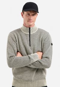Man met een zwarte pet en een beige gebreide trui met een kwart ritssluiting en geometrisch patroon, met zijn armen gekruist, neutrale achtergrond.