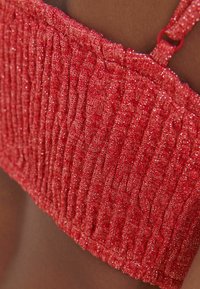 Punainen teksturoitu kangas, jossa on metallilankoja, esittää smokkiseosta ja ohuita säädettäviä olkaimia. Lähikuvassa korostuu materiaalin yksityiskohta.