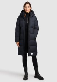 Lange zwarte pufferjas met capuchon en knoopsluiting. Beschikt over een doorstikte uitstraling, zijzakken en een logo-opdruk aan de onderkant. Ggedragen over zwarte kleding.