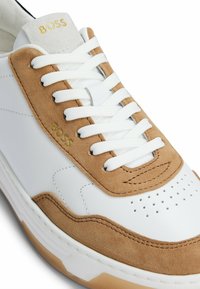 White leather sneaker with tan suede accents, perforated toe box, and white laces. Features a gray and gold logo on the tongue. Rubber sole.