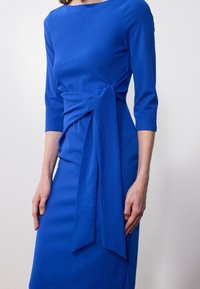 Robe bleue avec des manches trois-quarts, détail de nœud à la taille, tissu lisse et silhouette ajustée. Les accents incluent une ceinture en tissu.