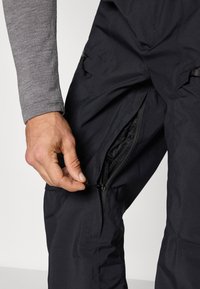 Calças pretas impermeáveis com fecho lateral e forro de rede. Close-up de uma mão segurando o puxador do fecho. O tecido é liso com acabamento mate.