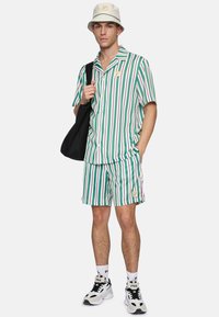 Randig kortärmad skjorta och matchande shorts i grön, vit och lila. Vit bucket hat med gröna detaljer, bärande en svart tote-väska.