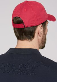 Rote Baumwollmütze mit verstellbarem Riemen, mit abgerundetem Hut und genähten Belüftungslöchern. Dunkles Poloshirt mit gestickten Details auf dem Rücken.